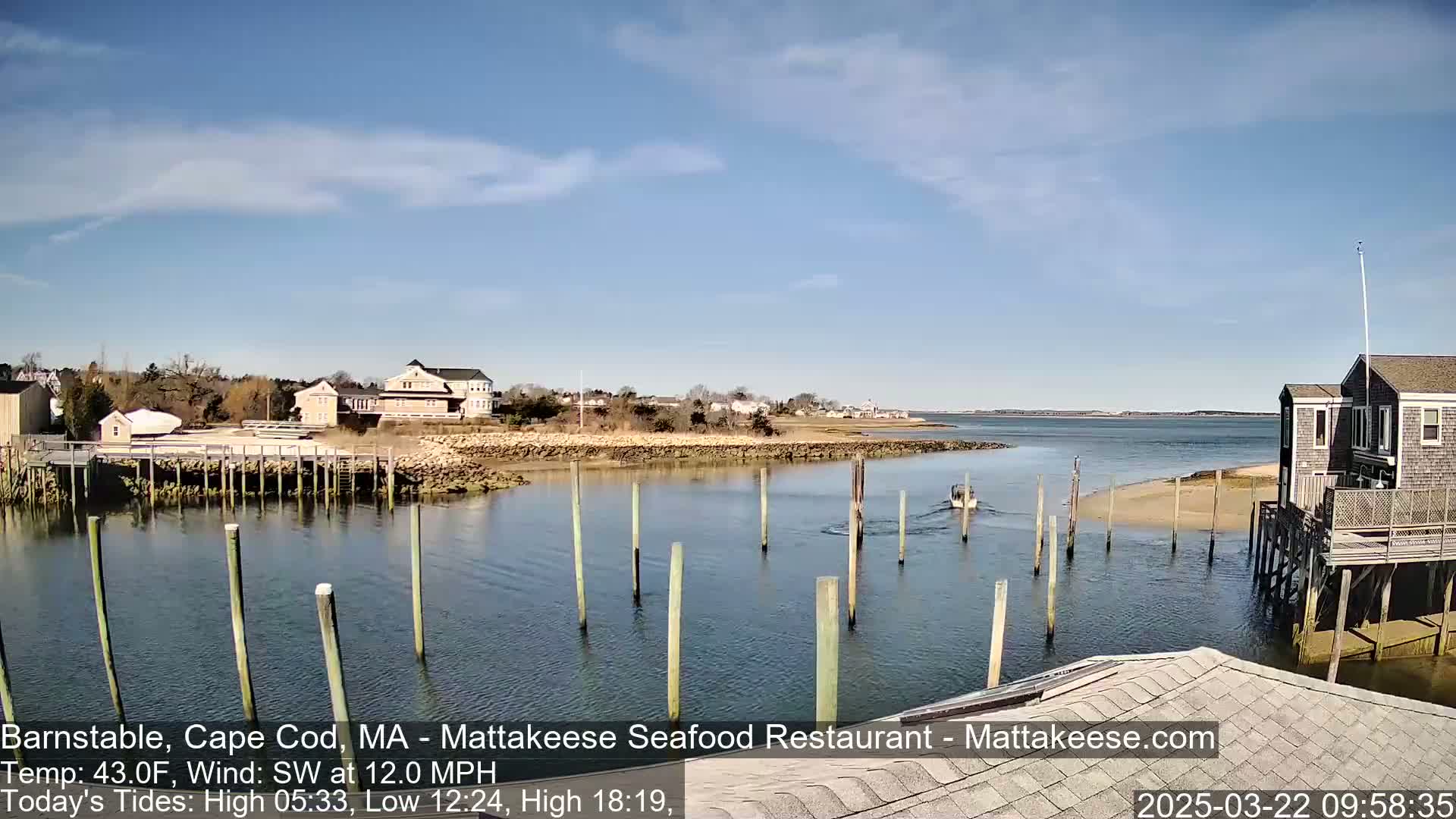 A calm, sunny day reveals a harbor scene with a small boat passing by wooden pilings, houses lining the shore, and a wide expanse of water under a mostly clear sky.
