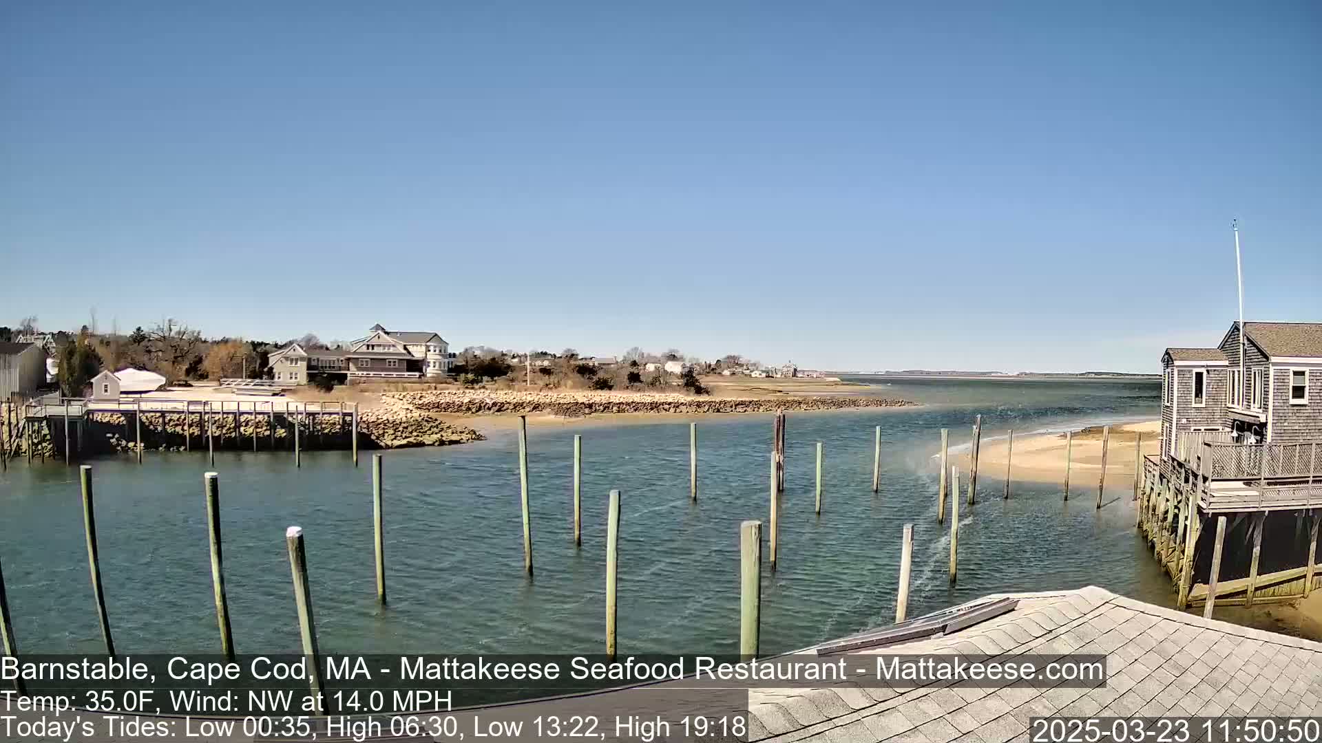 A clear, sunny day shows a calm waterway with several pilings in the water near a shoreline with houses and a small building on stilts.