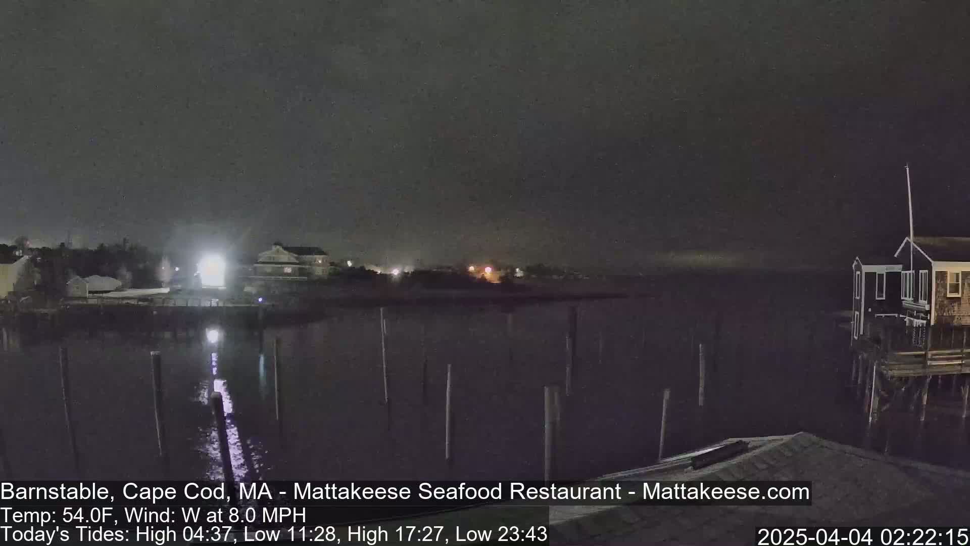 A nighttime view of a calm body of water with a few buildings and pilings visible across the water under a dark sky.
