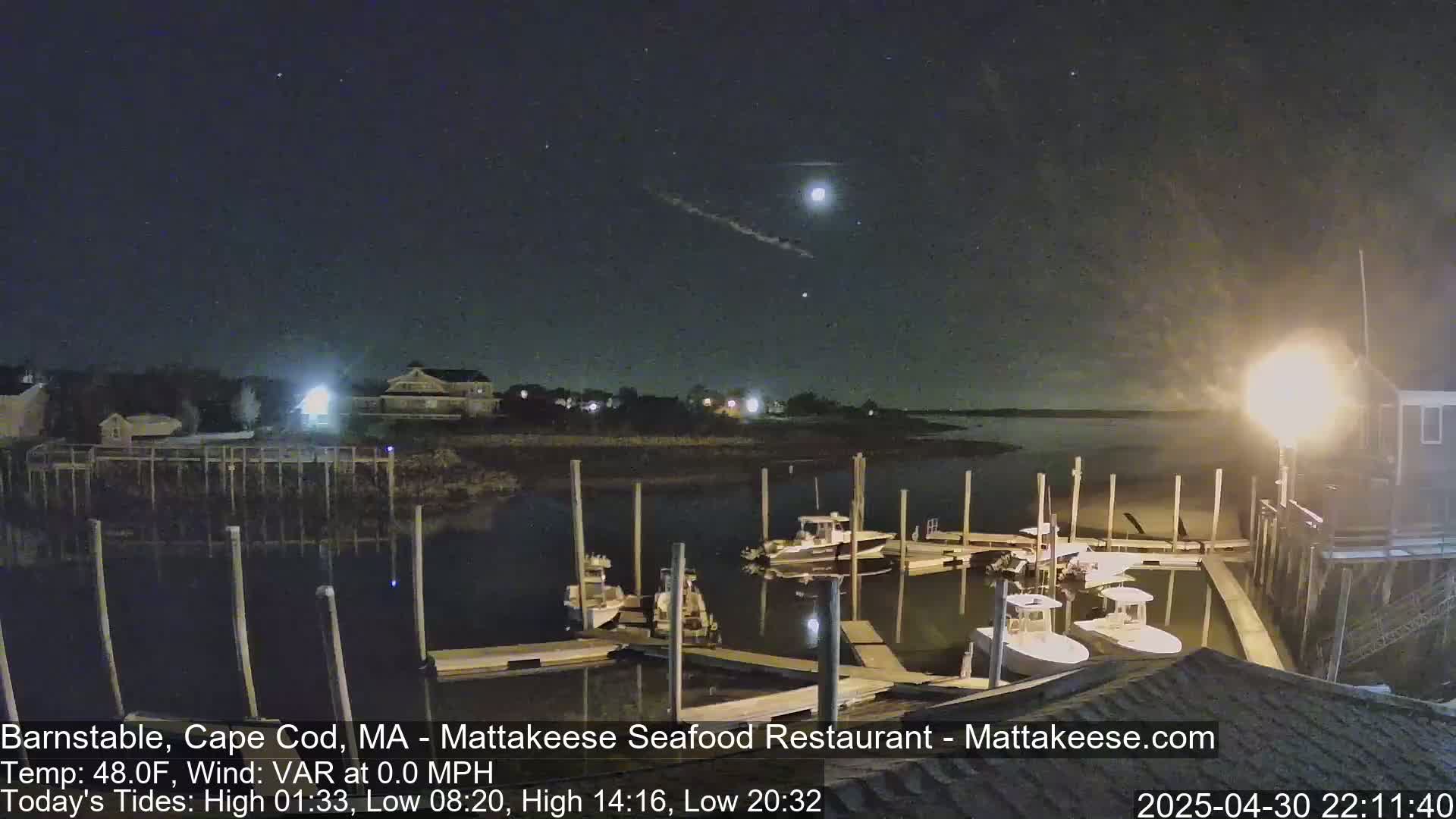 Several boats are docked at a marina at night under a partly cloudy sky, with a bright light illuminating the scene.