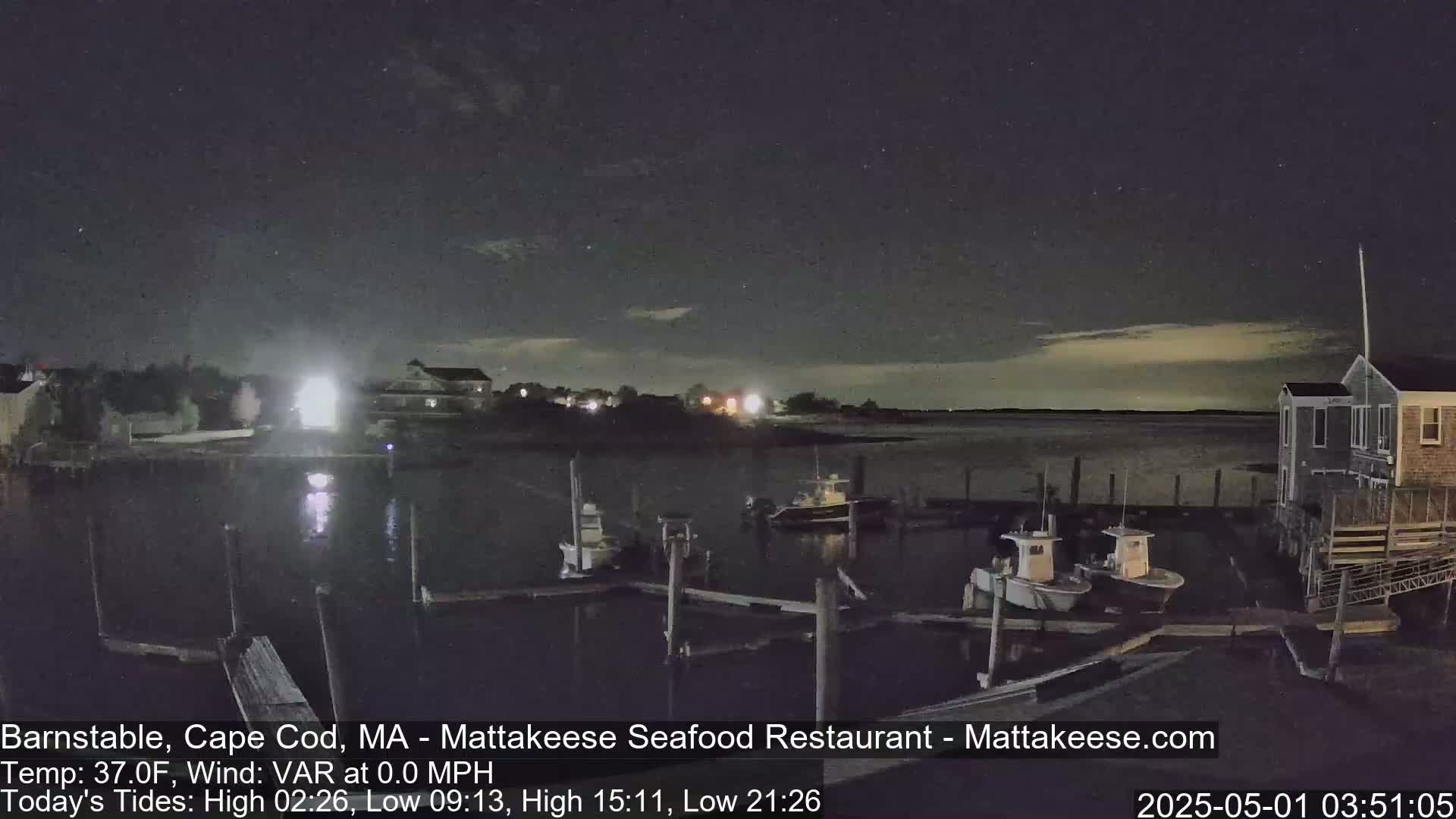 Under a dark, mostly clear night sky, several small boats are moored at a dock near a building, with calm water visible in the background.