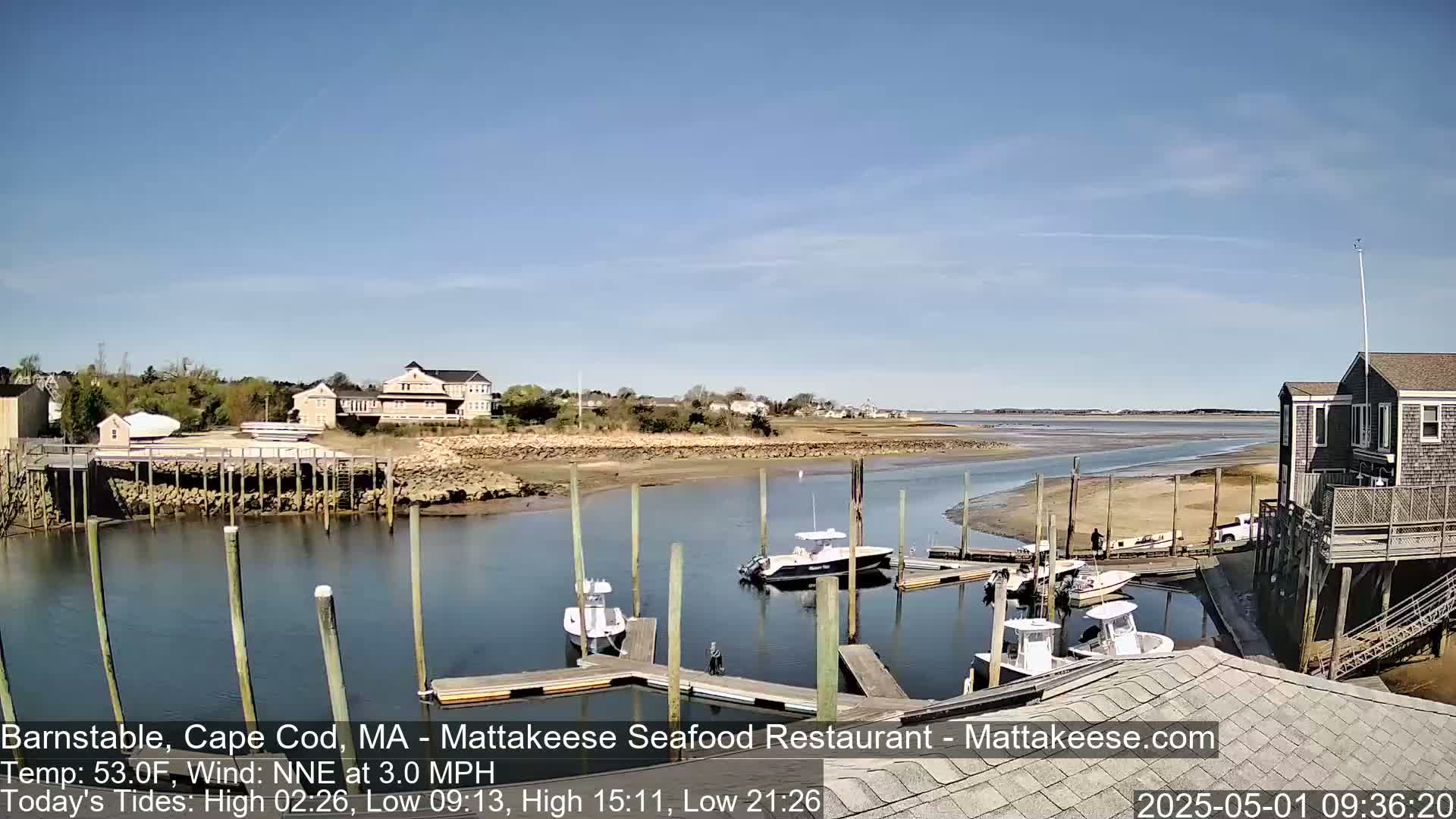 A calm, sunny day reveals a harbor scene with several boats docked at a marina, near houses and marshland.
