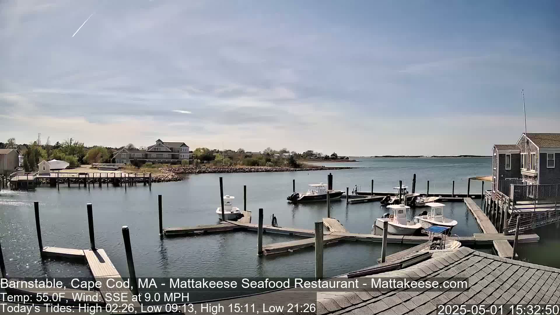 A calm, sunny day reveals a harbor filled with several docked boats, houses, and a small pier under a mostly clear sky with a faint contrail.
