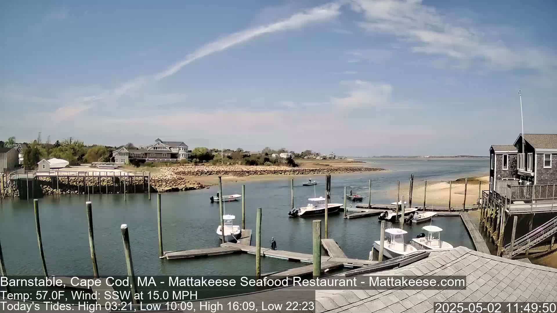 Several boats are docked at a marina on a sunny day with a light wind, overlooking a calm body of water and a shoreline with houses.