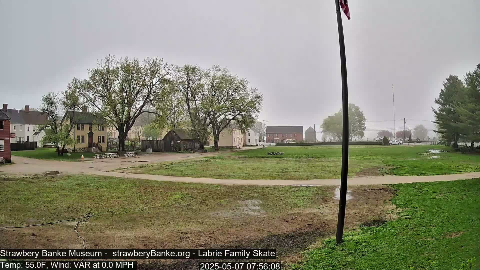 A grassy field with several historical-looking buildings and trees in the background on a foggy, overcast day.