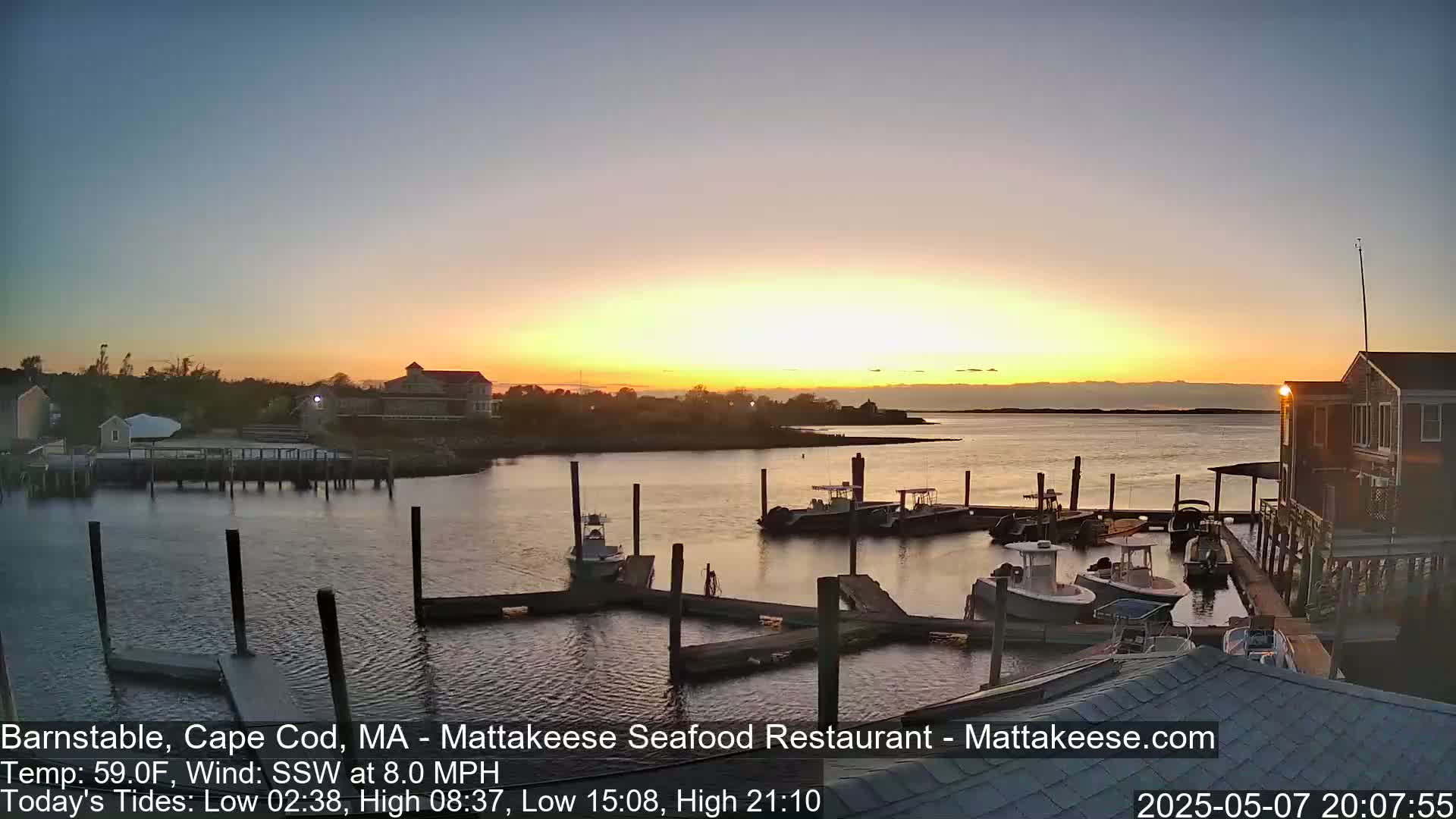 A calm harbor at sunset, with several boats docked along piers and buildings nearby.