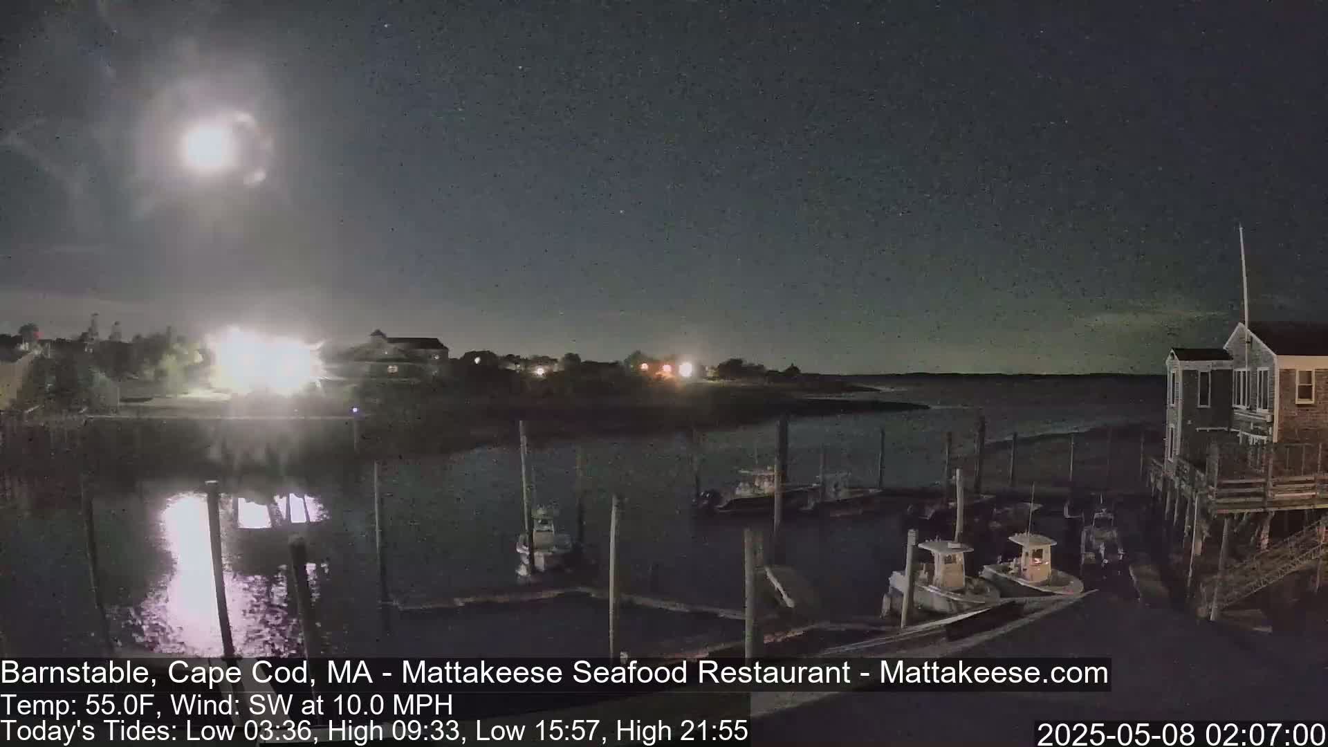 Under a bright moonlit night sky, several boats are moored at a dock near a waterfront building.