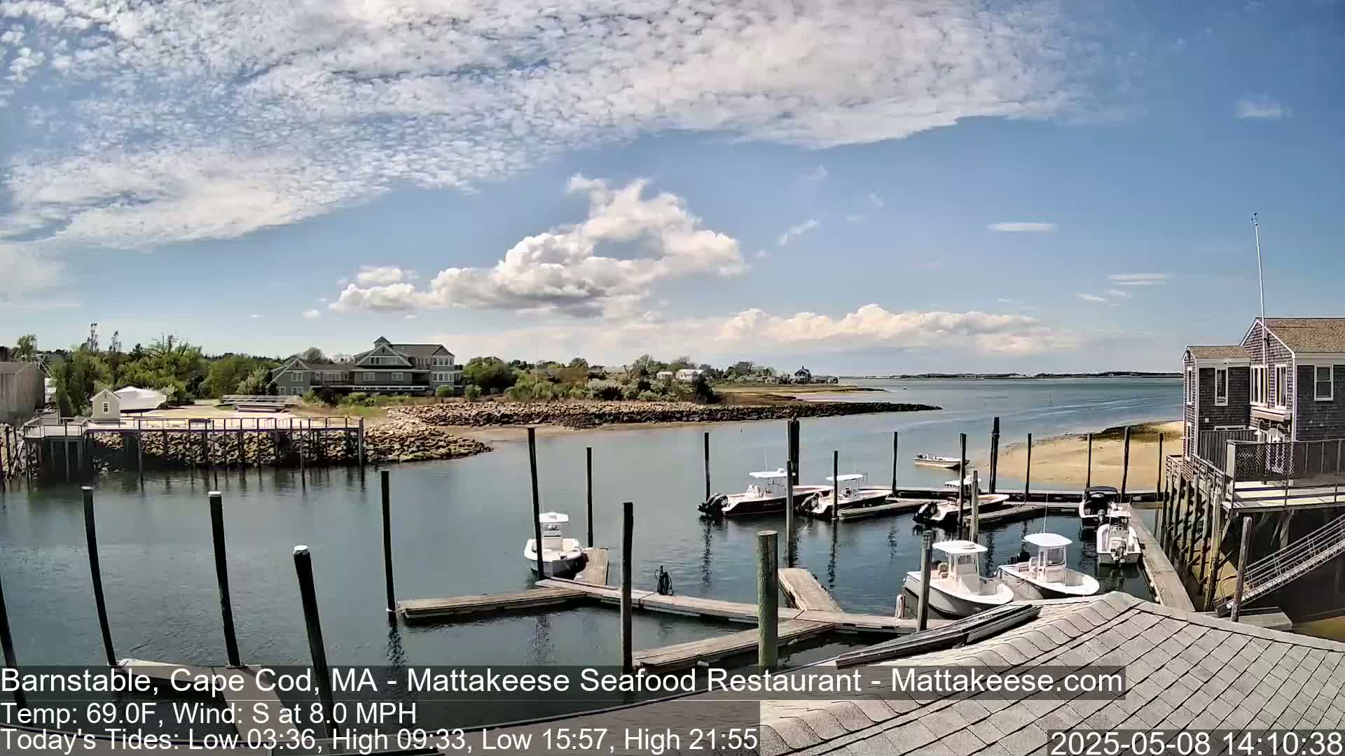 Several boats are moored at a marina on a partly cloudy day, with calm water and buildings visible in the background.