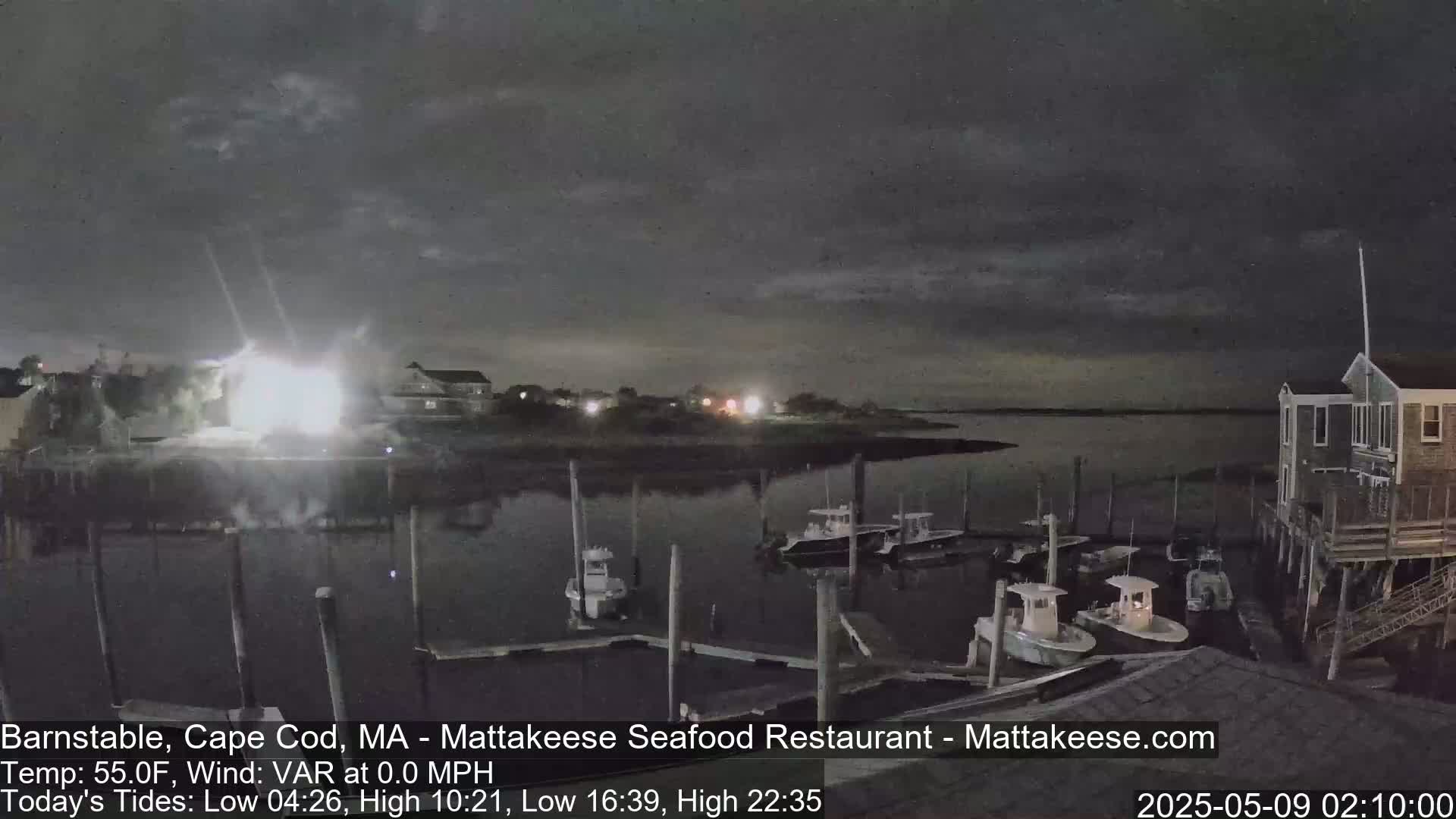 A nighttime scene shows a calm body of water with several small boats moored at a dock near buildings under a dark, cloudy sky.