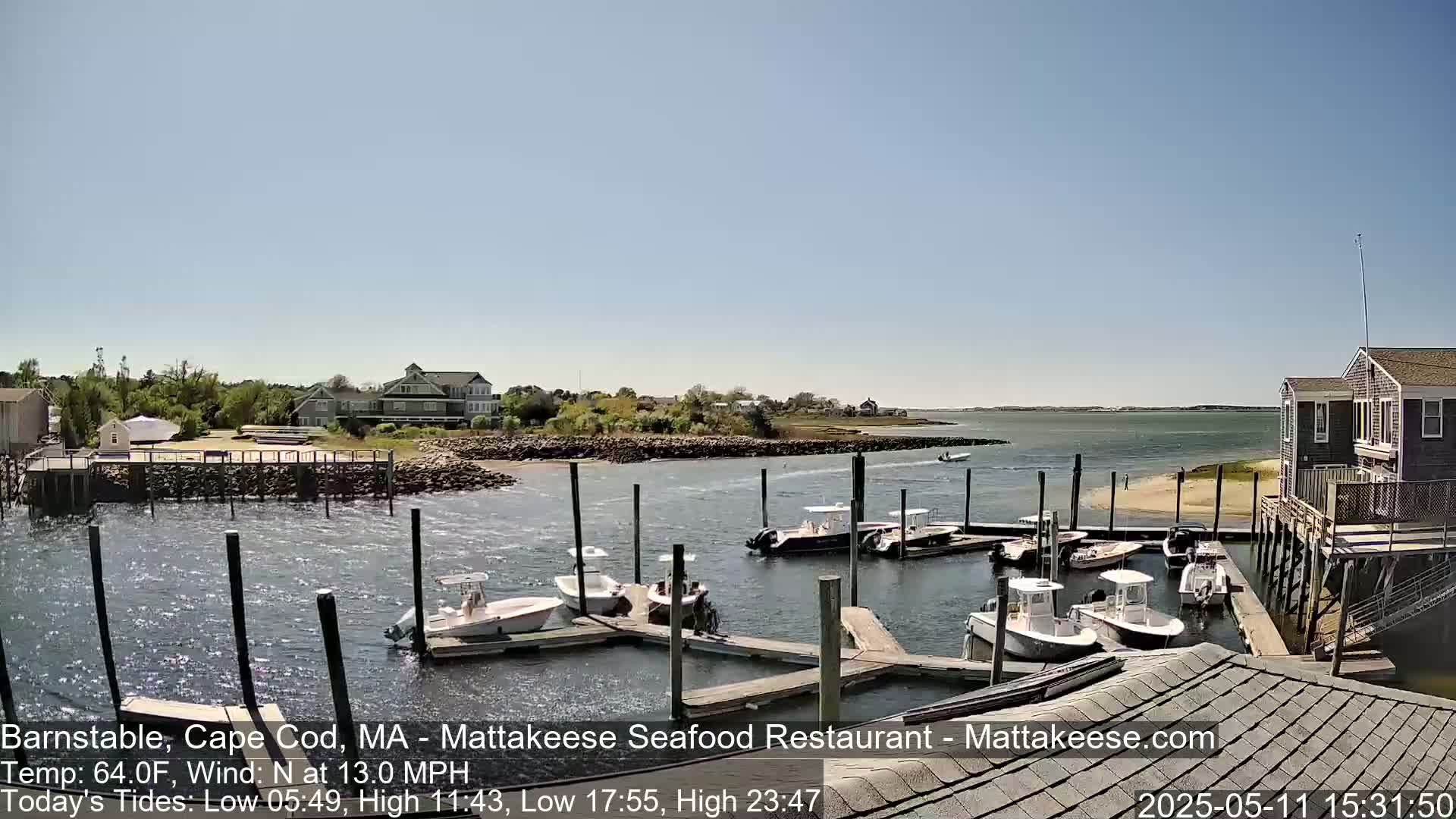 Several boats are docked at a marina on a sunny day with a north wind of 13 mph, overlooking a calm body of water and a shoreline with houses.