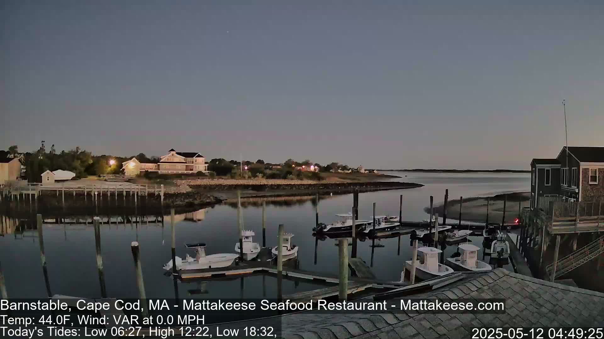 Several boats are moored at a calm marina at twilight, with buildings and trees visible on the shoreline.