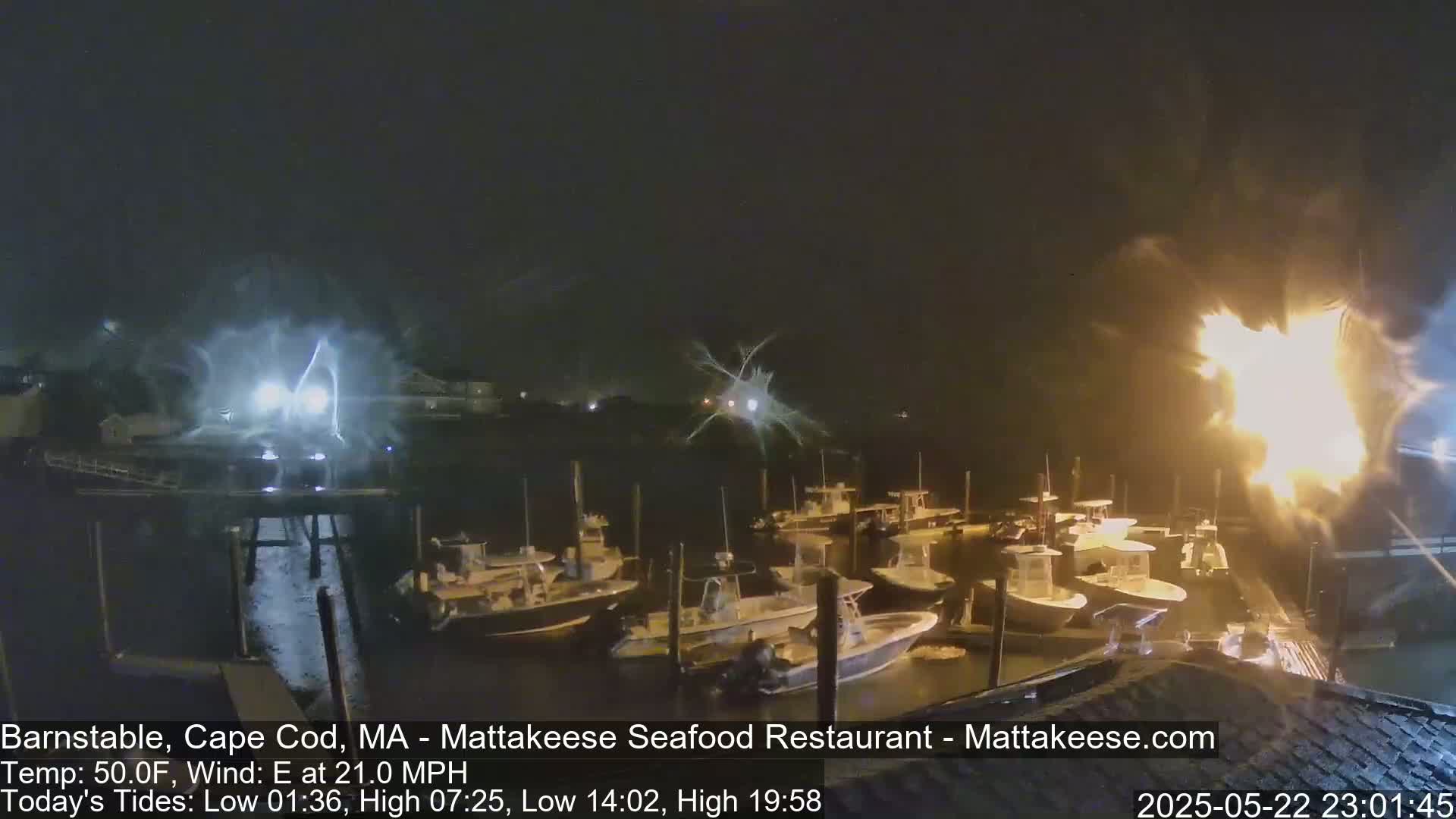 A nighttime view of numerous boats docked at a marina under a dark sky with bright flashes of light in the distance and a strong east wind.