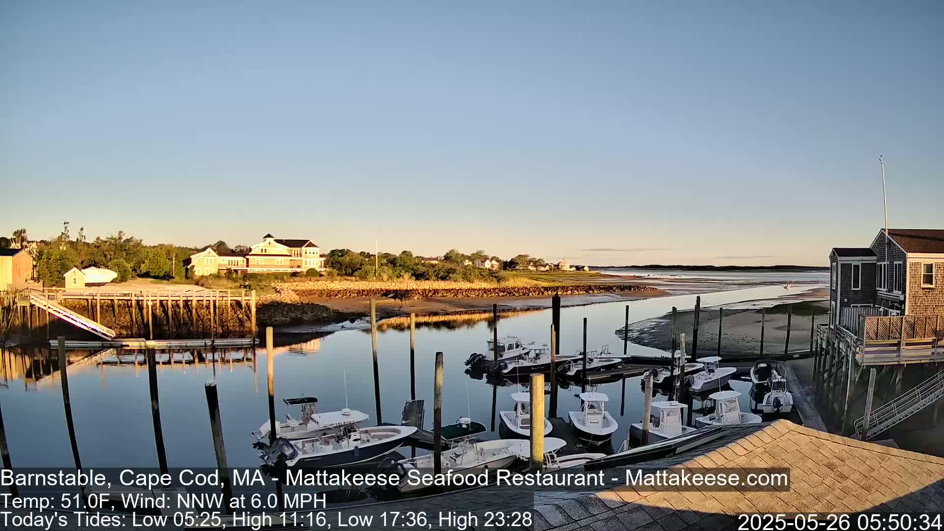 Several boats are docked at a calm harbor on a clear, sunny morning, with buildings and houses visible along the shoreline.