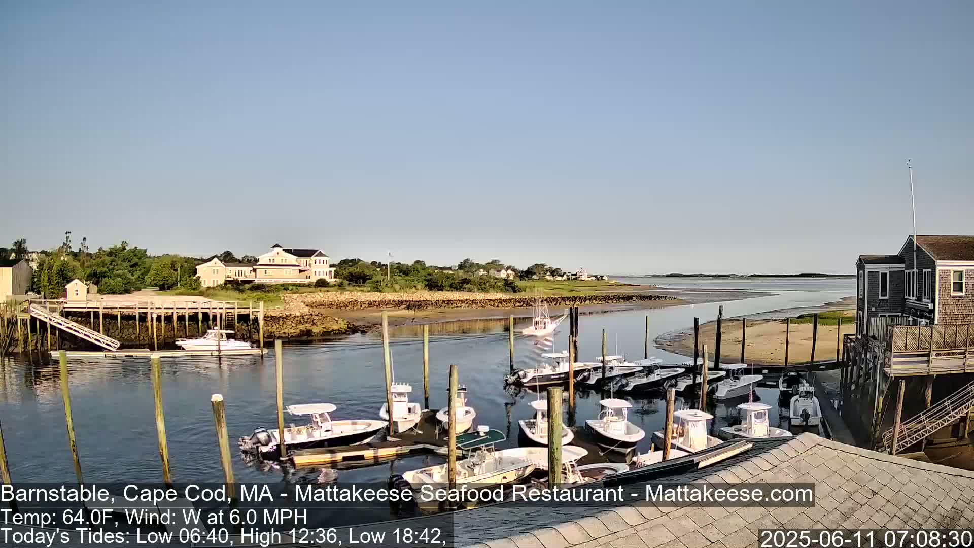 A calm harbor with numerous small motorboats docked, a larger boat entering the harbor, and houses and trees visible on the shoreline under a clear sky.