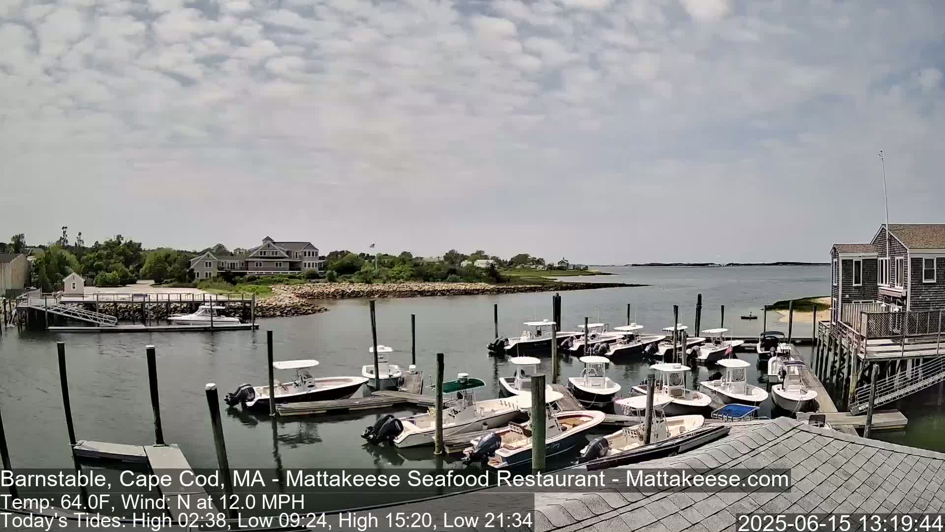 A calm, partly cloudy day shows many boats docked at a marina next to a waterfront building, with additional houses visible across the water.