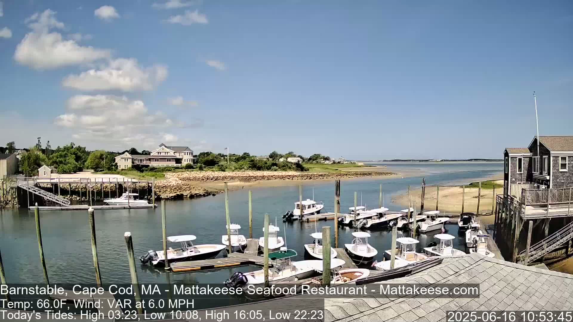 A sunny day shows a calm harbor with numerous boats docked at a wooden pier, near buildings and a sandy shoreline.