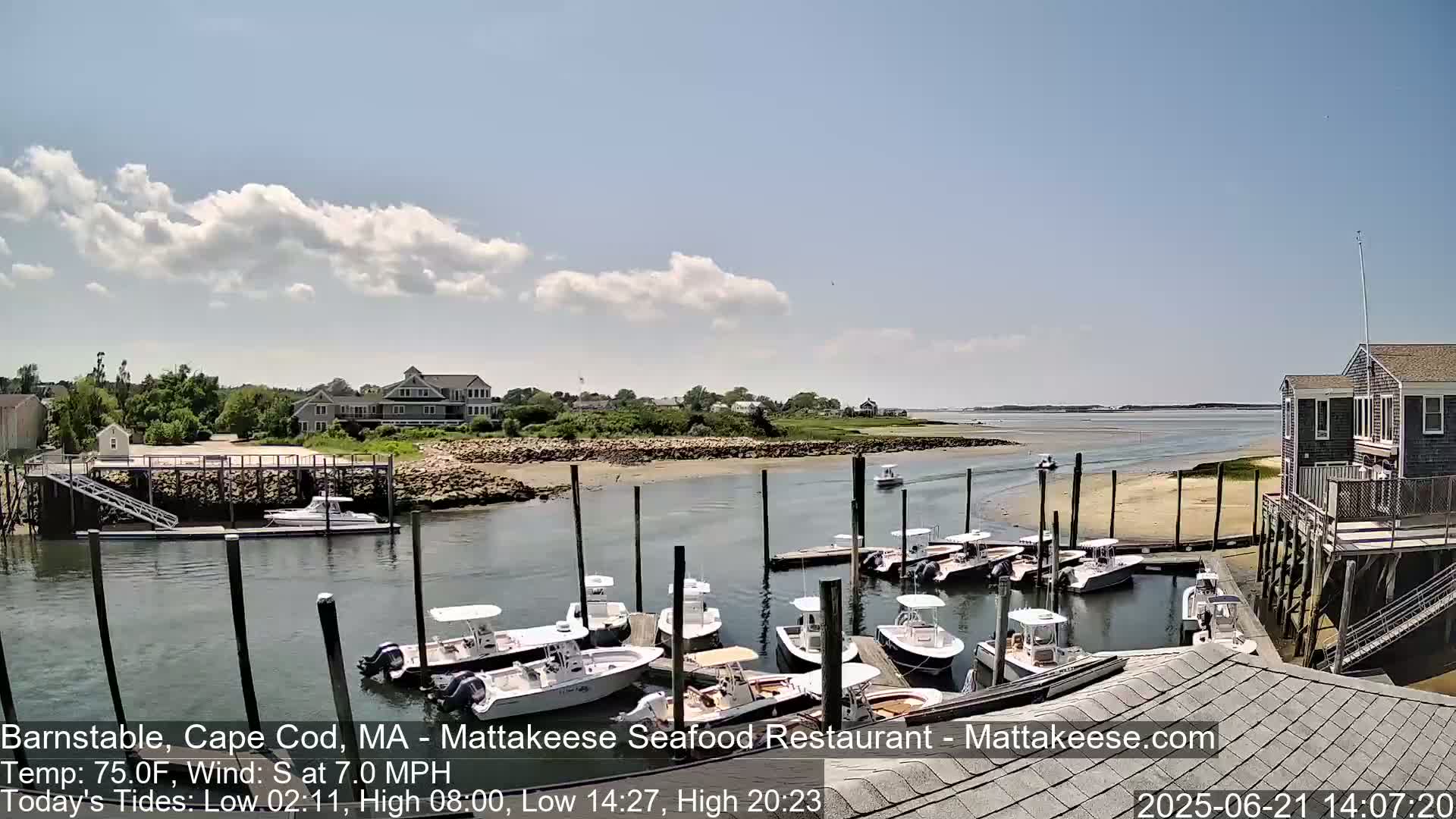 A sunny day shows a harbor with numerous boats docked, alongside a building on pilings and other buildings in the background.
