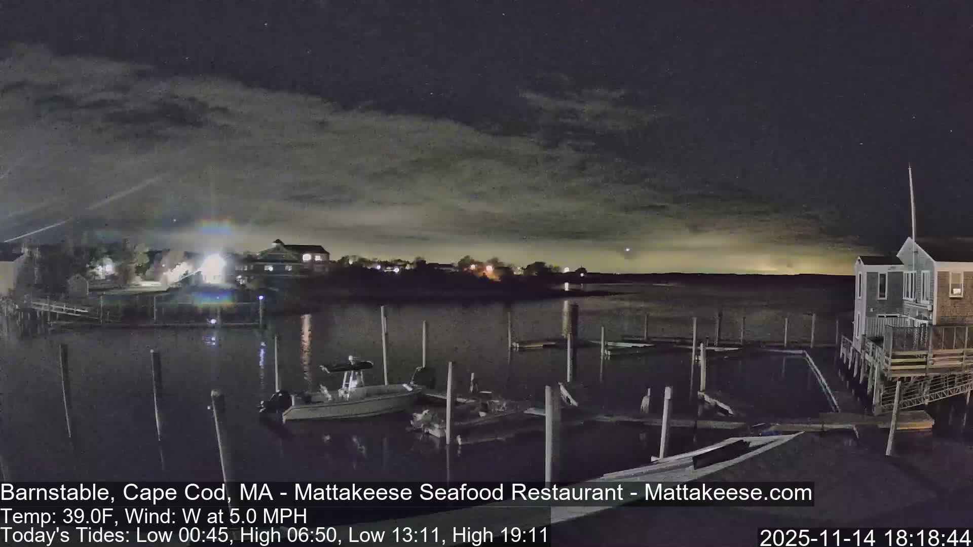 A serene night view of a harbor features boats docked in the foreground, illuminated buildings across the water, and a large boathouse on the right, all under a partly cloudy sky with calm water reflecting the distant lights.