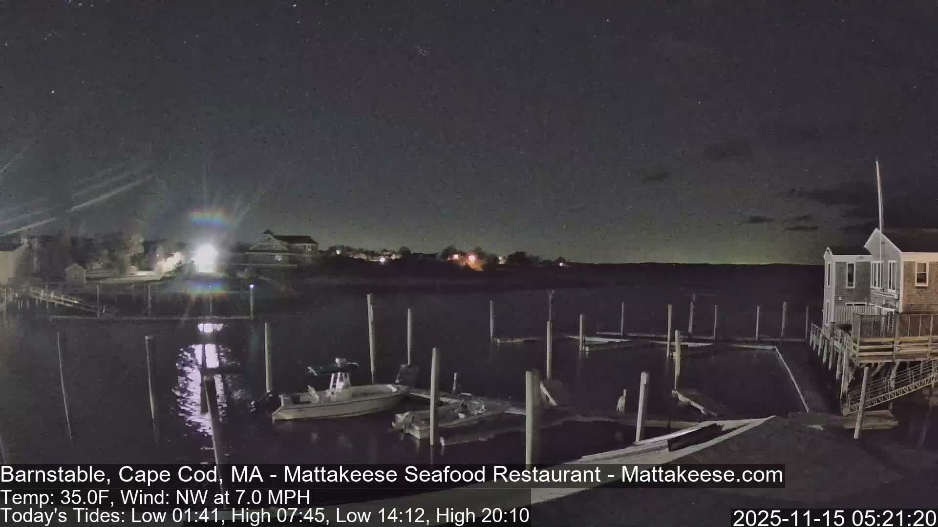 A clear, starry night reveals a peaceful marina with boats moored at various wooden pilings and buildings along the distant shore.