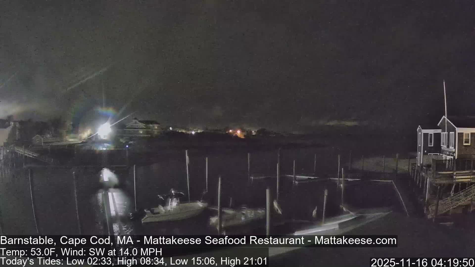 The image shows a dark, cloudy night over a marina with several boats moored at docks, illuminated by various artificial lights, including a bright source on the left producing a strong lens flare.