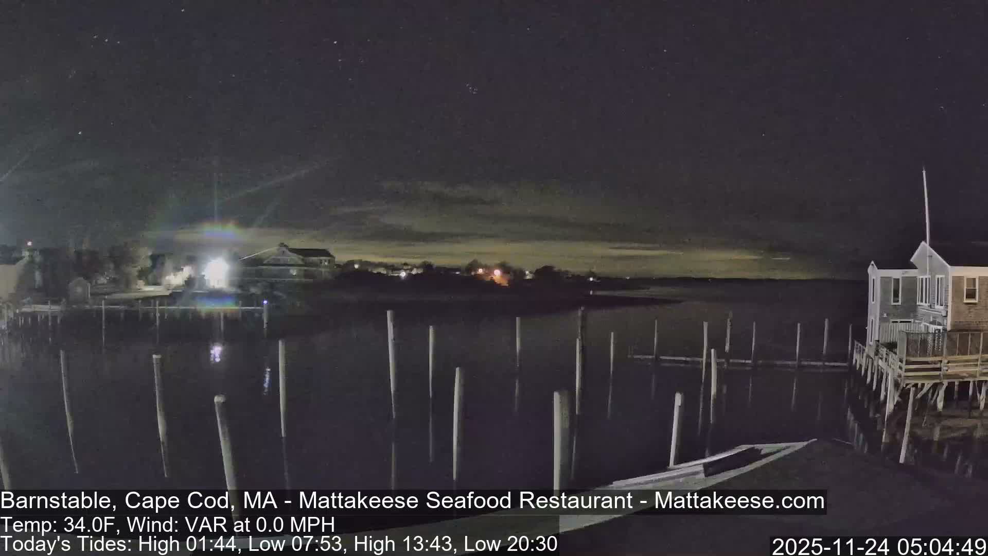 This nighttime image depicts a tranquil waterfront with numerous pilings in calm water reflecting bright lights from shore buildings, all beneath a dark sky faintly revealing stars and some distant cloud cover.
