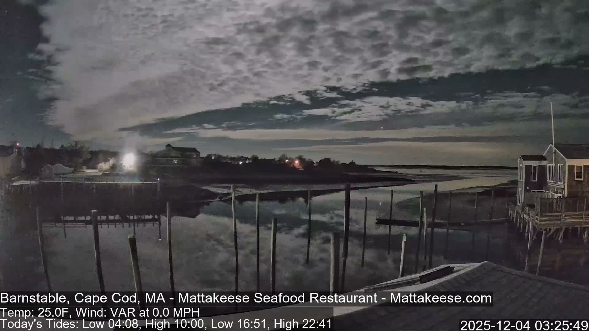 A cloudy night reveals a tranquil tidal inlet featuring numerous pilings reflecting the sky and distant shore lights, with a small shingled building on stilts visible to the right.