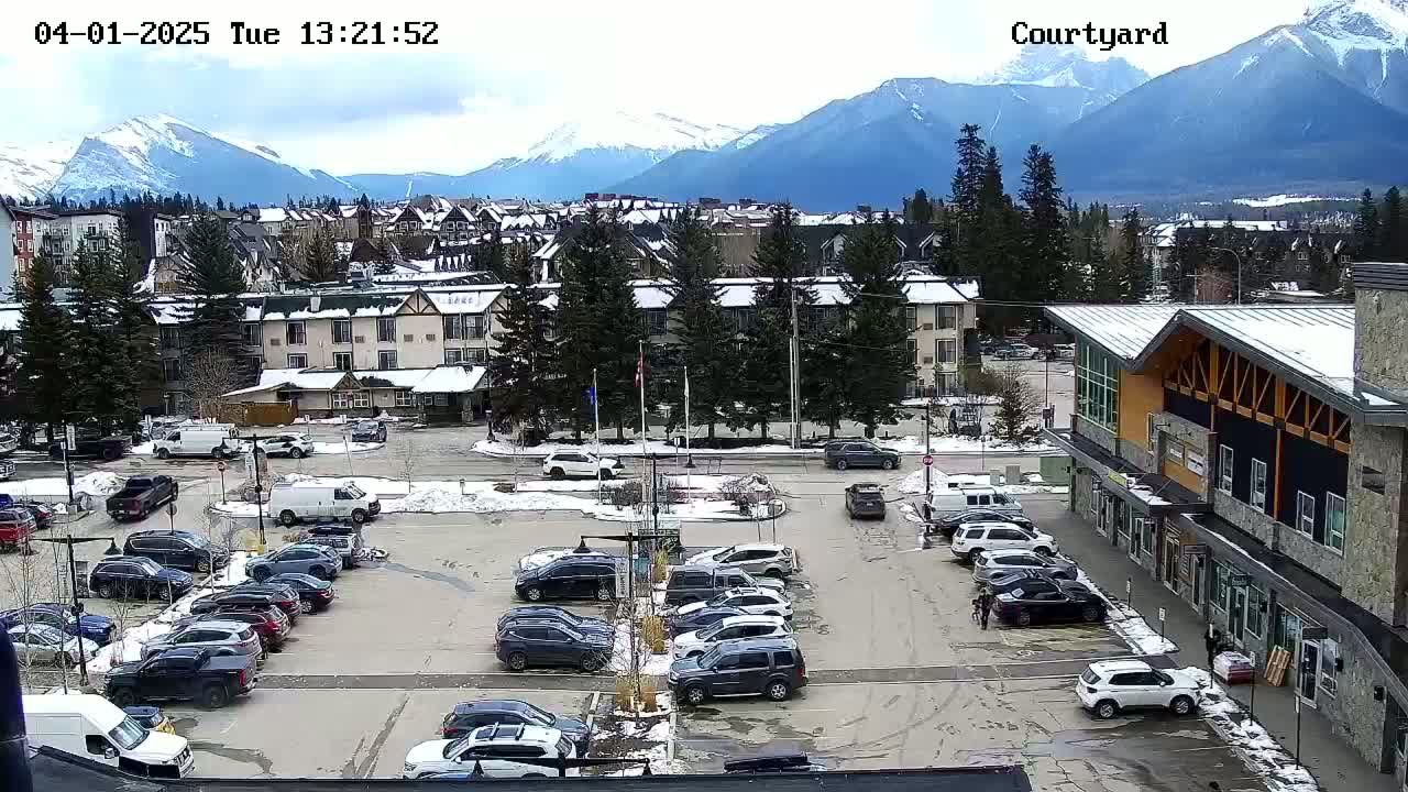 A partly cloudy day reveals a snow-dusted parking lot filled with cars, situated in front of buildings and overlooking a mountain range.