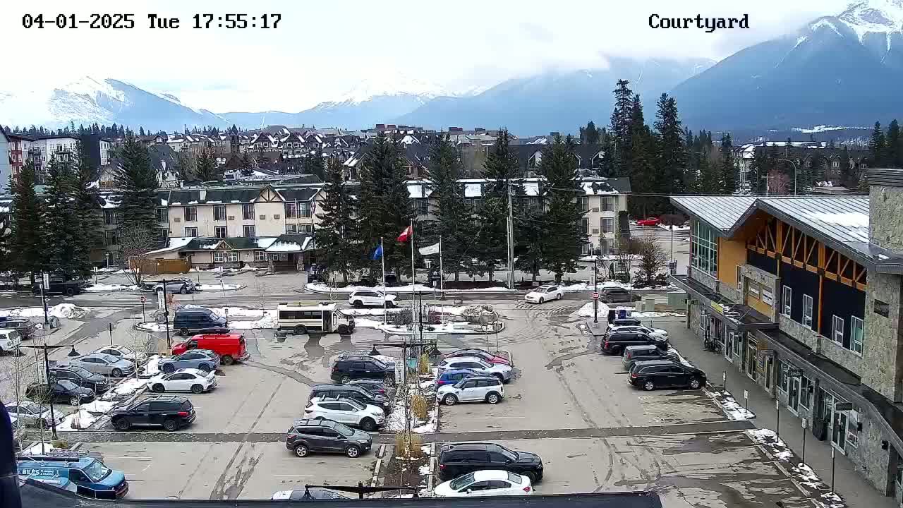 A parking lot with numerous cars is situated in front of buildings, with a mountain range visible in the background under a cloudy sky.