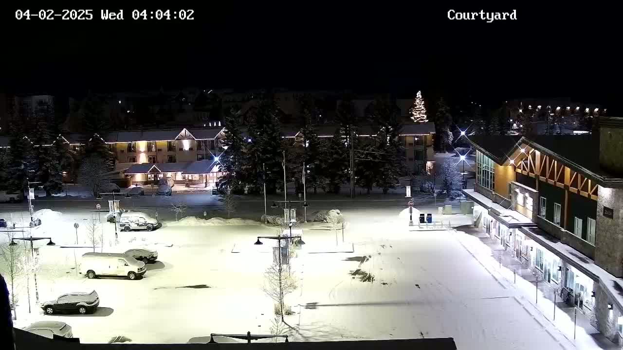 A snow-covered parking lot at night, illuminated by lights, with a hotel and other buildings in the background.