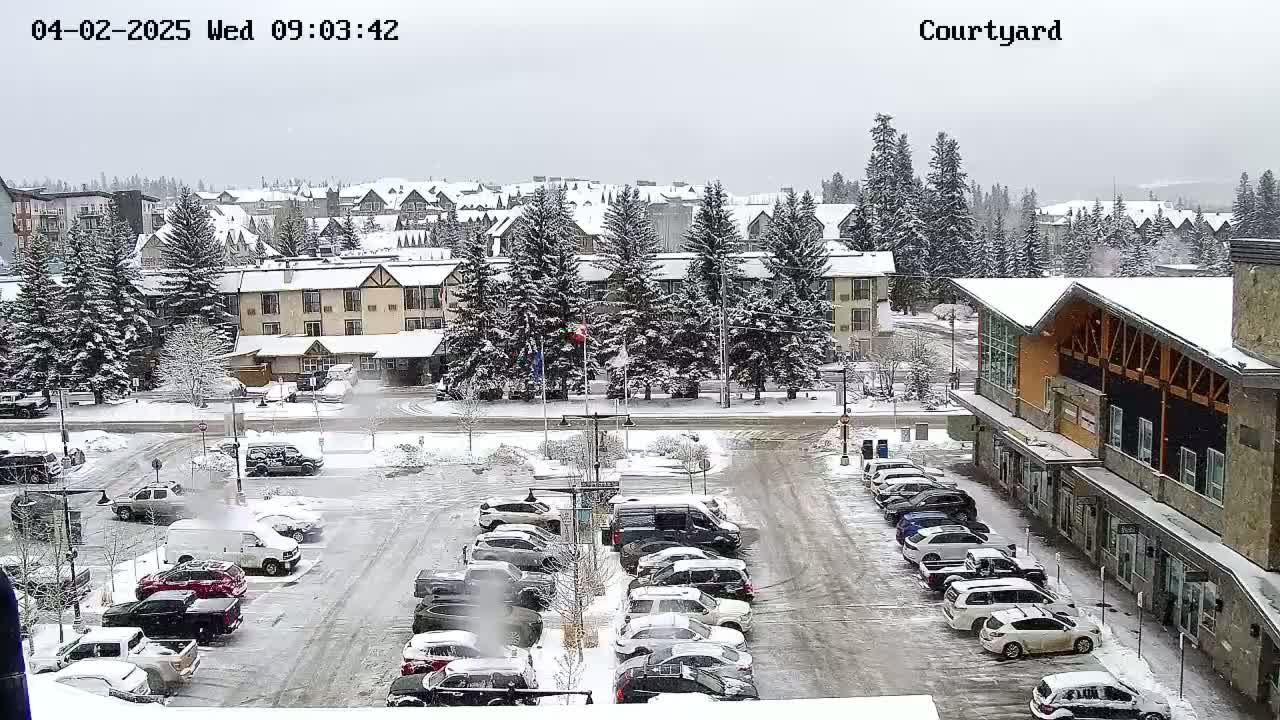 A snow-covered parking lot with cars is in front of a multi-story building and other snow-covered buildings, under a gray, overcast sky.