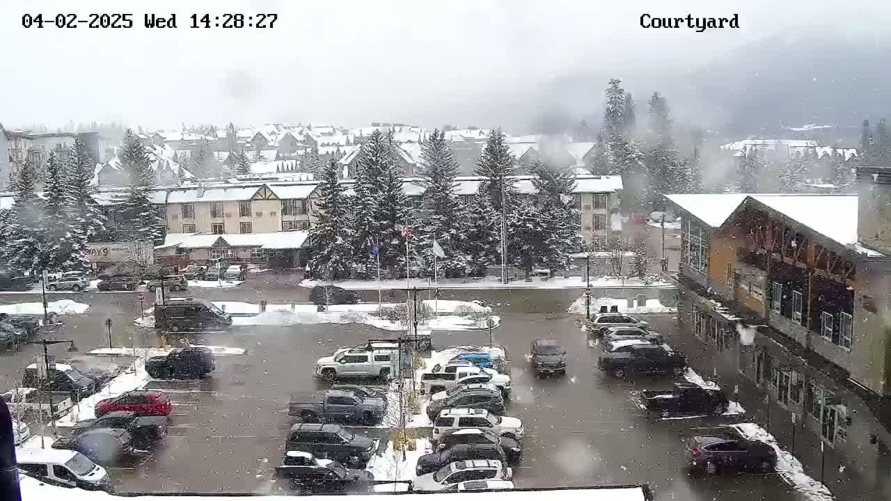 A snow-covered parking lot and town are viewed from above, with snow falling.
