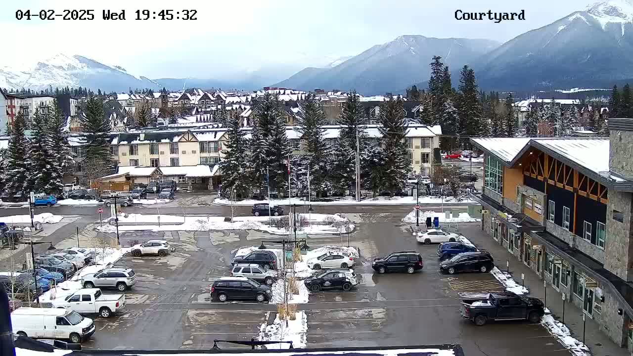A snow-covered parking lot with cars and trucks, surrounded by buildings and evergreen trees, sits in front of a mountain range under a cloudy sky.