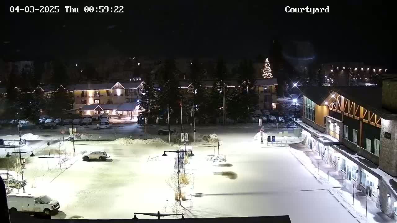 A snow-covered courtyard at night, illuminated by artificial lights, shows a hotel and other buildings with some vehicles parked in the lot.