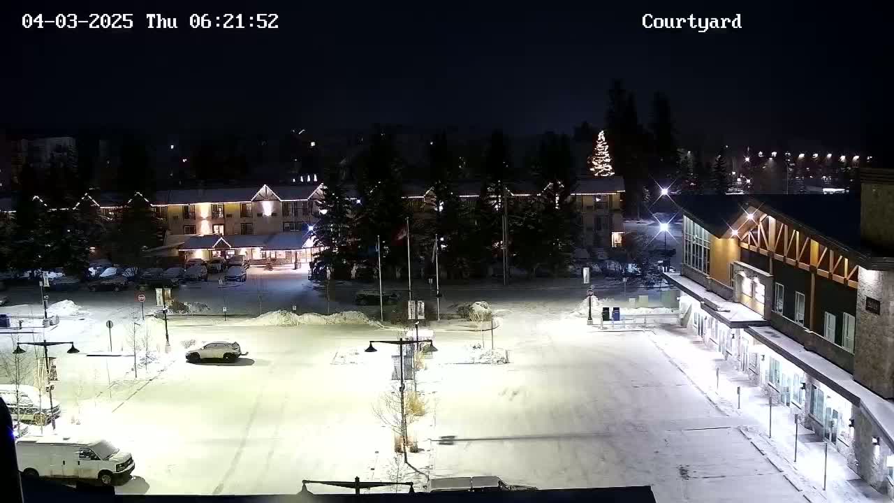 A snow-covered parking lot at night, illuminated by lights, with a hotel and other buildings in the background.