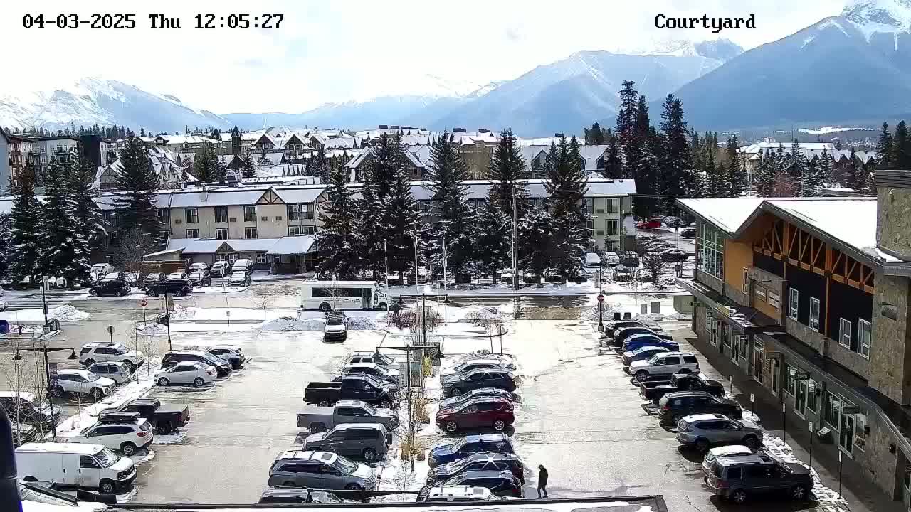A snow-covered parking lot with cars, surrounded by buildings and evergreens, sits in front of a mountain range under a mostly sunny sky.