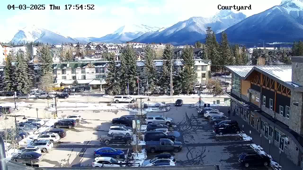 A snow-dusted parking lot with numerous vehicles is situated in front of buildings, with a backdrop of snow-capped mountains under a mostly sunny sky.