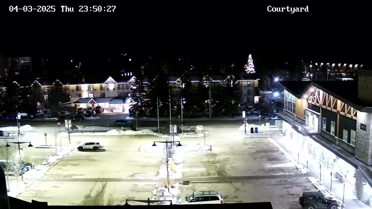 A nighttime, snow-covered parking lot is illuminated, showing several cars parked and a few buildings in the background.