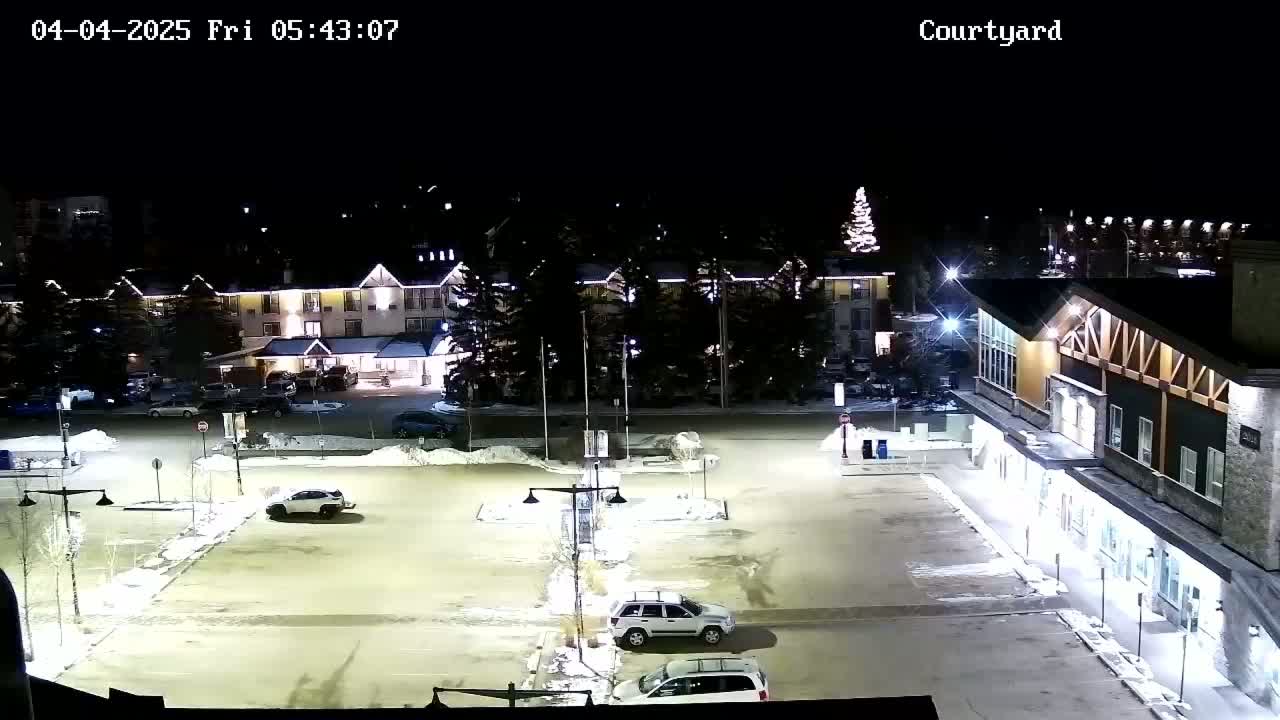 A snow-covered parking lot at night, illuminated by artificial lights, is surrounded by buildings and trees.