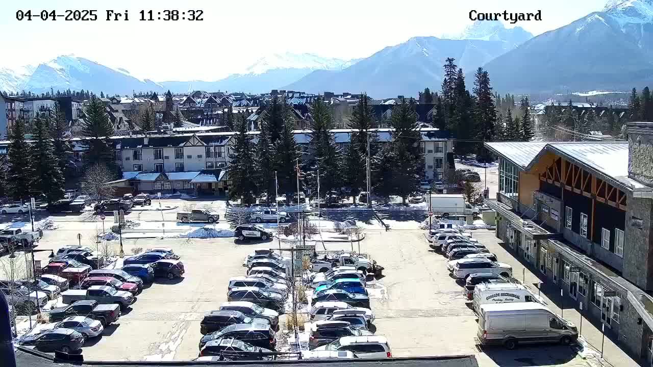 A sunny day reveals a parking lot filled with cars in front of buildings and trees, with snow-capped mountains in the background.