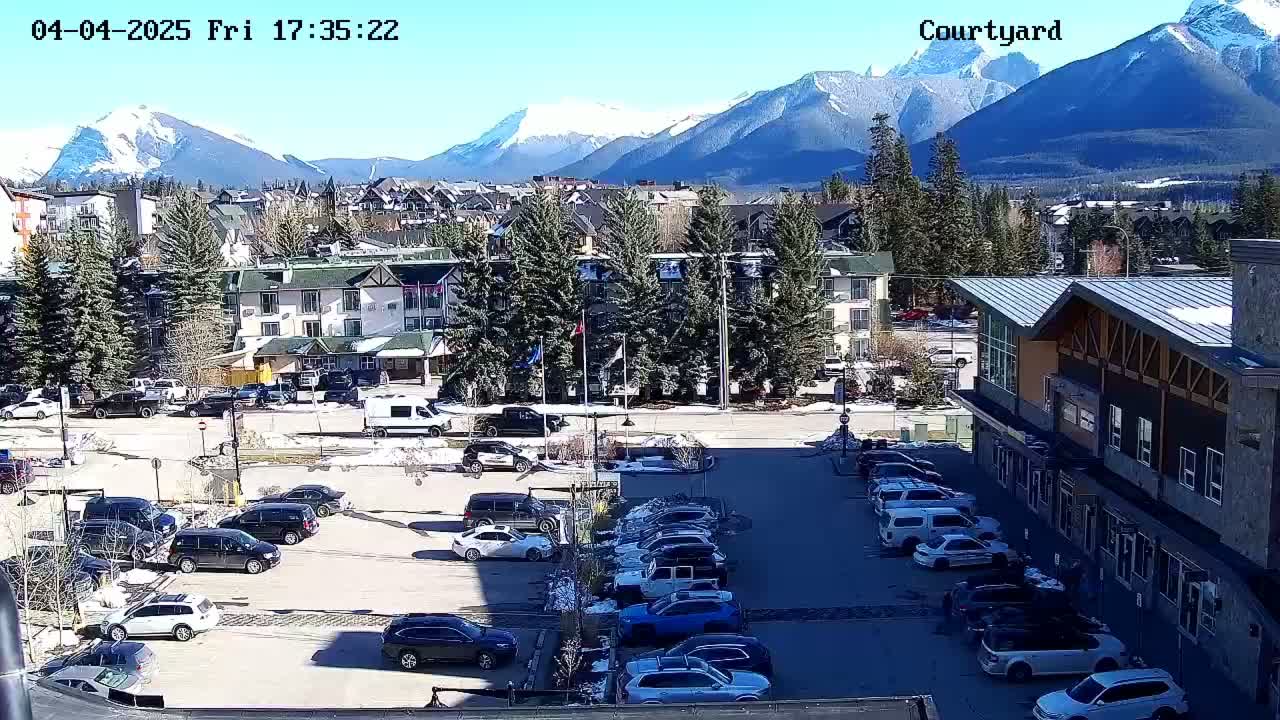 A sunny parking lot with several cars is situated in front of buildings, with snow-capped mountains visible in the background.