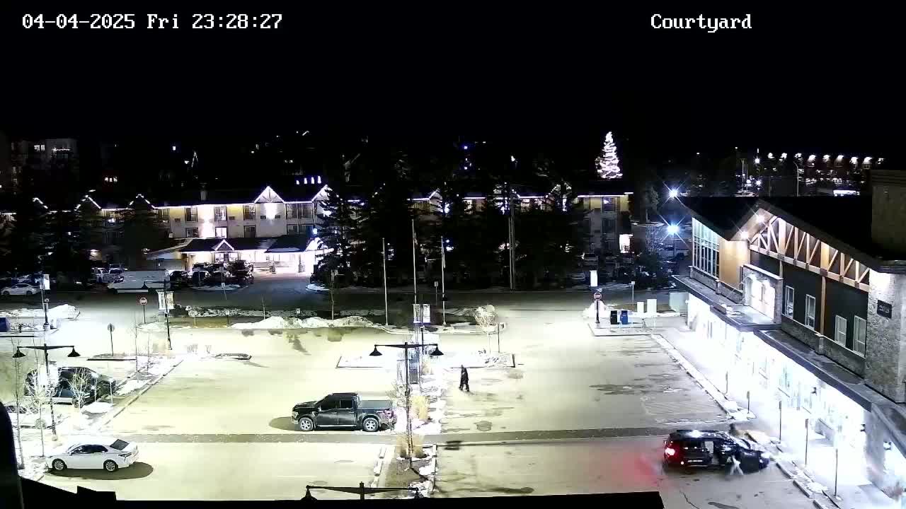 A nighttime view of a snow-covered parking lot between buildings, with several vehicles and a person walking.