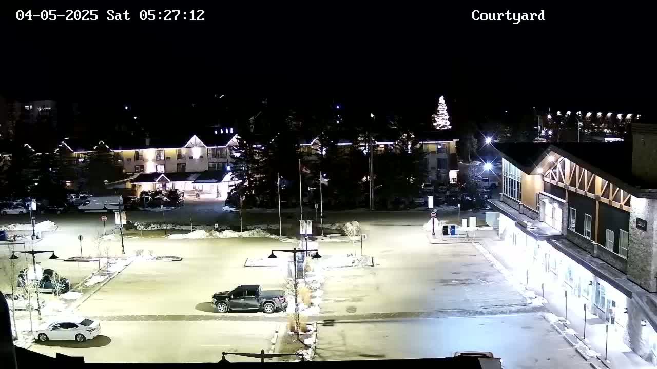 A nighttime view of a snow-covered parking lot with several vehicles, illuminated buildings, and sparsely spaced trees.