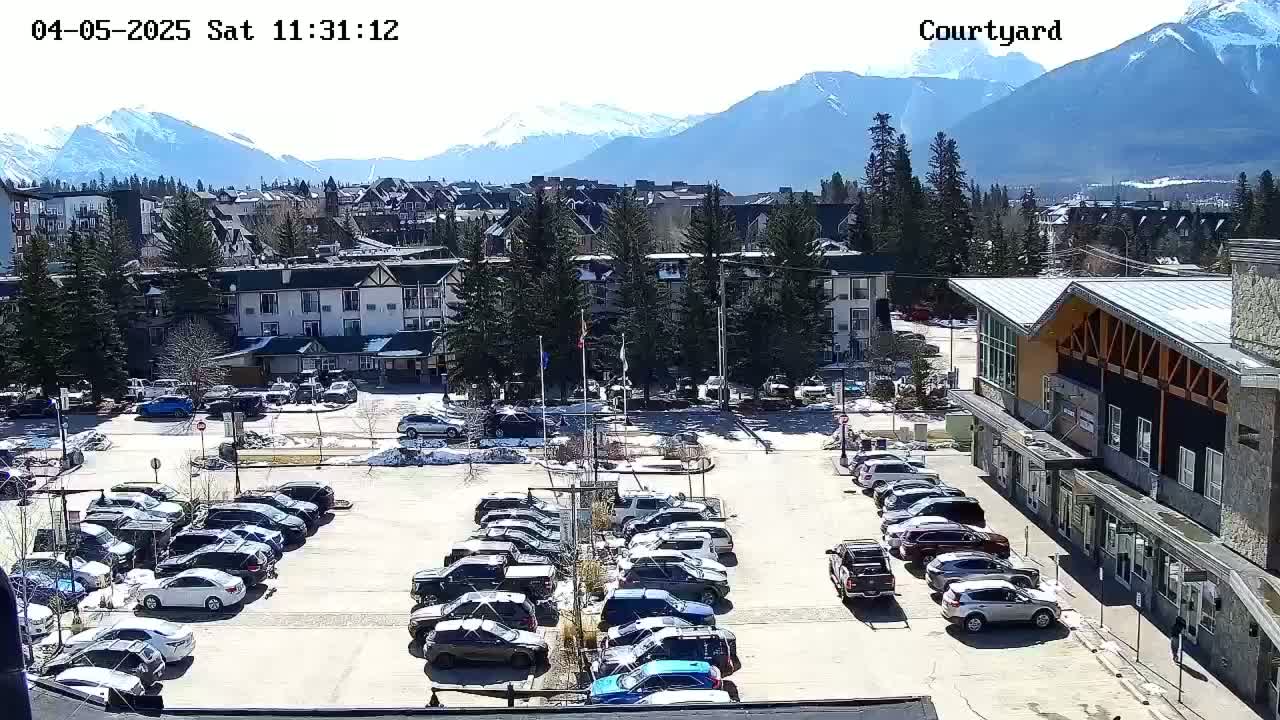 A sunny day reveals a parking lot full of cars in front of buildings nestled at the base of a snow-capped mountain range.
