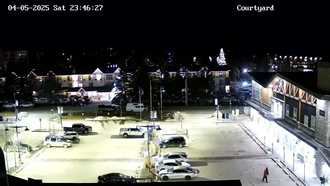 A nighttime, snow-dusted parking lot with several cars and buildings, including a hotel, lit up against a dark sky.
