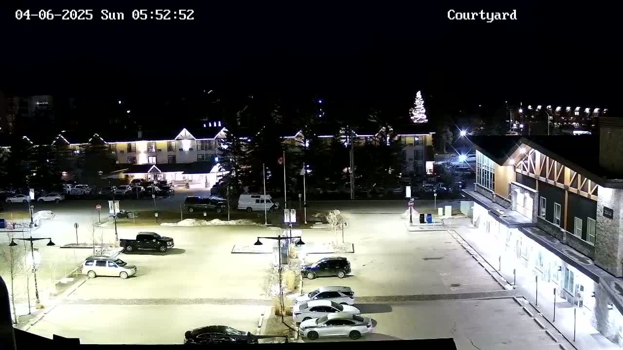 A nighttime, snow-covered parking lot with several cars, a large hotel, and a building with a long, lit facade.