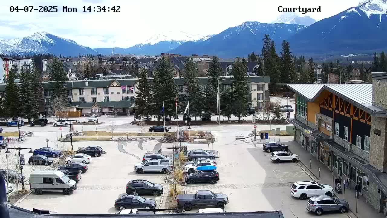 A parking lot with numerous cars is situated in front of a multi-story building, with a mountain range in the background on a partly cloudy day.