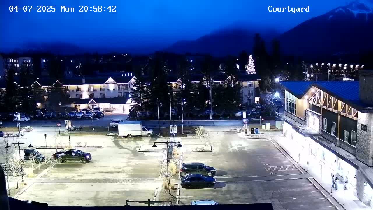 A nighttime view of a parking lot with several vehicles, illuminated by streetlights, situated in front of a hotel and other buildings, with mountains in the background under a dark, clear sky.