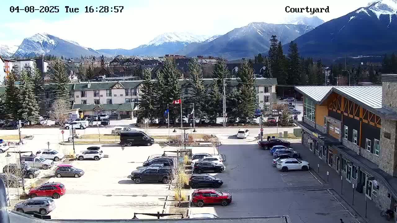 A parking lot with numerous vehicles is situated in front of a multi-story building, with snow-capped mountains and a sunny sky visible in the background.