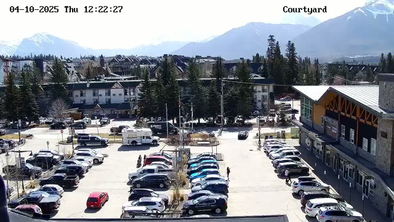 A sunny parking lot filled with cars is situated in front of a multi-story building and other structures, with a mountain range visible in the background.
