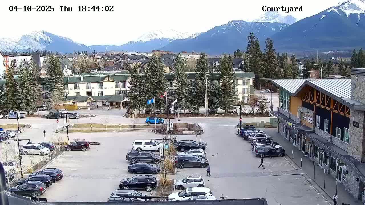 A parking lot with cars and a few pedestrians is situated in front of buildings, with snow-capped mountains visible in the background under a mostly clear sky.